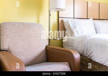 Das Schlafzimmer war in der Nähe von einem Sessel mit Armsitz und weißen Kissen mit Lampe und Bettlaken auf dem Kopfteil des Matratzenbettes in einem modernen Motelzimmer mit niemand Stockfoto