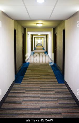 Das moderne Inn-Hotel Motel verfügt über einen leeren langen Korridor mit Teppichboden und Lampen, die eine vertikale Innenansicht der Gebäudearchitektur beleuchten Stockfoto