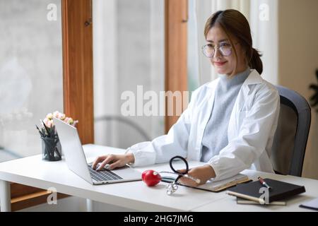 Professionelle Ärztin, die an ihrem Schreibtisch sitzt und an ihrem Laptop arbeitet. Stockfoto