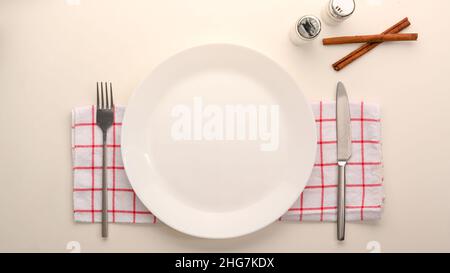 Leere weiße Keramikplatte Mockup mit Besteck auf rot-weißem Küchentuch auf Esstisch. Flach Stockfoto