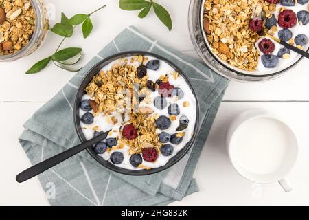 Frühstück mit Müsli und frischen Heidelbeeren und Himbeeren mit Joghurt in Schüsseln auf einem weißen Holztisch.Draufsicht.gesundes Esskonzept Stockfoto