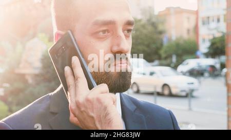 Porträt eines Geschäftsmannes in diplomatischem Anzug, der am Telefon telefoniert und mit einem entfernten Kollegen über die Marketingstrategie diskutiert. Manager, der draußen vor dem Büro des Startup-Gebäudes steht Stockfoto