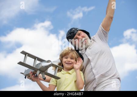 Lustige Großvater und Sohn spielen mit Holzflugzeug vor Sommerhimmel Hintergrund. Kind Junge mit Träumen vom Fliegen oder Reisen. Generationsfamilie. Stockfoto