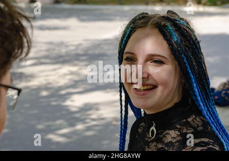 Ein junges heterosexuelles Paar plaudert, während es auf einer Bank in einem Stadtpark sitzt. Eine Frau mit langen blauen Dreadlocks. Der Mann mit Brille. Selektiv Stockfoto