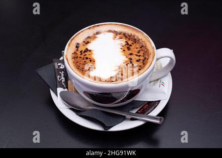 Kaffee – ein Cappuccino mit Schokoladenüberzug, der in einer Teetasse mit Untertasse, Zuckerbeutel und Teelöffel serviert wird Stockfoto
