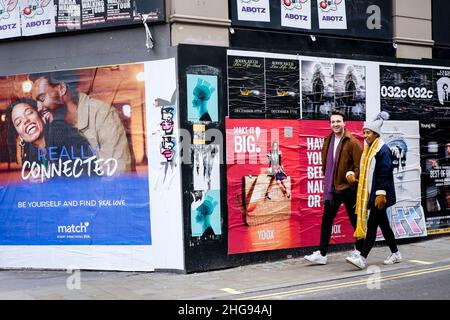 Lächelnd, geht das junge Paar an einer mit Fliegenplakaten bedeckten Wand vorbei, von der eines eine Dating-Website anwirbt. London, Großbritannien. Stockfoto