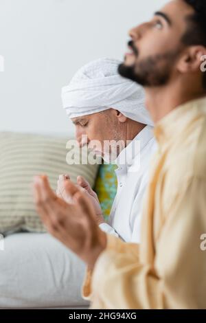 Seitenansicht eines reifen Mannes, der mit geschlossenen Augen in der Nähe eines verschwommenen arabischen Sohnes zu Hause betet Stockfoto