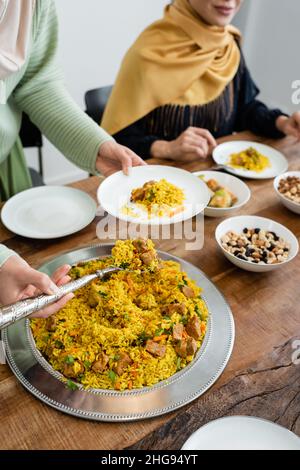 Beschnittene Ansicht einer jungen muslimischen Frau, die zu Hause Pilaf serviert, fast unscharf Mutter und Essen Stockfoto
