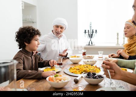 muslimischer Mann mittleren Alters, der während eines Familienessens mit einer interracial Familie auf einen arabischen Enkel zeigt Stockfoto