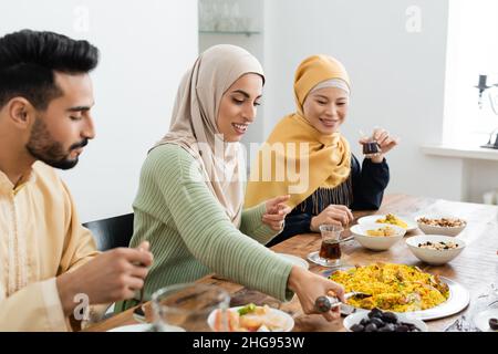 muslimische Frau, die Pilaf in der Nähe der asiatischen Mutter und des verschwommenen Ehemanns serviert Stockfoto