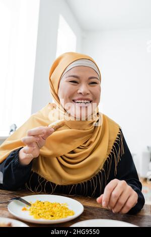 Fröhliche asiatische Frau in Hijab hält süße Baklava in der Nähe Pilaf auf dem Teller Stockfoto