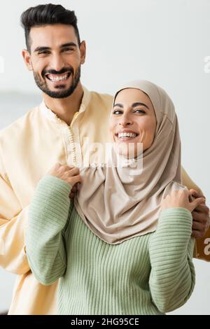 Fröhlicher arabischer Mann umarmt junge Frau in Hijab, während er die Kamera anschaut Stockfoto