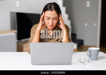 Unglückliche Freiberuflerin mit gemischter Rasse hat eine Frist mit einer Remote-Arbeit. Müde Frau hat keine Zeit, pünktlich zu Ende zu gehen, sie sitzt im Heimbüro und berührt den Kopf mit der Hand Stockfoto