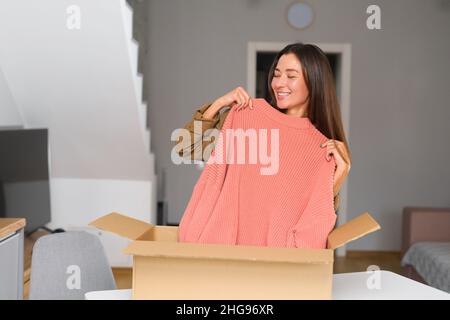 Zufriedene stilvolle asiatische Frau öffnet Paketkarton Box, Auspacken empfangene Geschenk, Gefühl neugierig auf bestellte Artikel aus einem Online-Shop, weibliche Kunden glücklich mit schneller Lieferung und Versand Stockfoto