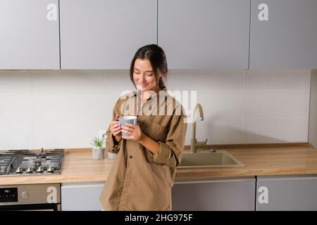 Finden Sie Gleichgewicht im Leben. Friedliche und ruhige Asiatin verbringt den faulen Morgen zu Hause, steht mit Becher in den Händen in der Küche und genießt Kaffee Stockfoto