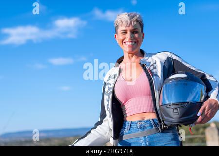Die junge, hübsche blonde Frau mit kurzen Haaren, die als Biker mit einer Lederjacke und ihrem Schutzhelm gekleidet ist, lacht beim Blick auf die Kamera Stockfoto