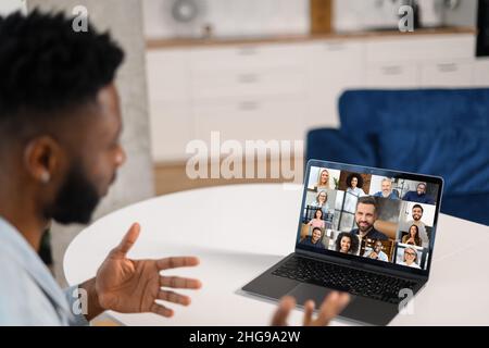 Remote-Meeting per Videoanruf. Multirassischer Mann, der in der Küche sitzt und die App auf dem Laptop für die Online-Kommunikation mit einer Gruppe verschiedener multirassischer Menschen verwendet. Rückansicht Stockfoto
