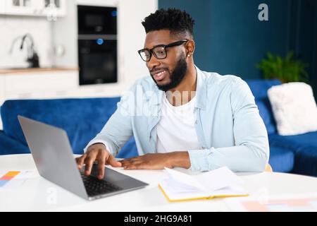 Selbstbewusster, konzentrierter, multirassischer Freiberufler, der im Heimbüro am Laptop arbeitet, während er am Schreibtisch sitzt und den Computer benutzt. Brunette Mann tippen auf der Tastatur, Blick auf den Bildschirm, genießen Remote-Arbeit Stockfoto