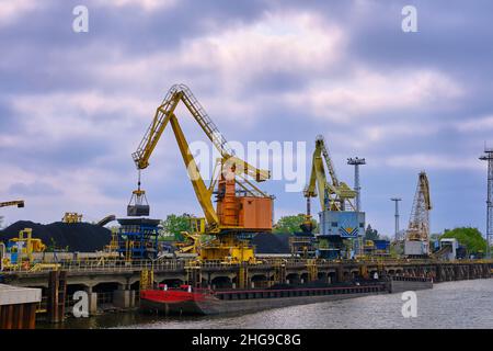 Flussthafenkrane mit Clamshell oder Greifer laden Kohle zu Fluss ziehen Boote oder Lastkähne am Pier an bewölktem Tag festgemacht. Energie, Mineralien, Umwelt Stockfoto