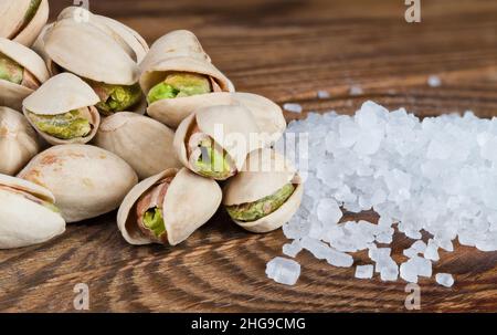 Nahaufnahme von gerösteten Pistazien-Nusshaufen und Tafelsalz auf einem hölzernen Hintergrund. Pistacia Vera. Gesalzene grüne Früchte in offenen, hart gespaltenen, halbierten beigefarbenen Schalen. Stockfoto