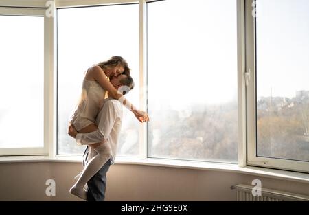 Sorgloses junges Paar steht am Fenster und umarmt und küsst sich im Blick auf die Stadt. Konzept der Flitterwochen im Urlaub. Stockfoto