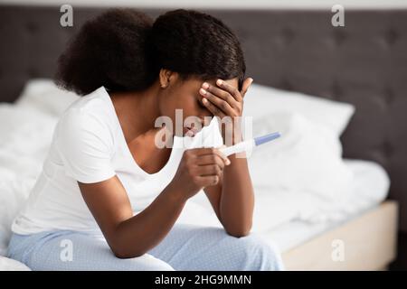 Verärgerte afroamerikanische Frau, die mit einem Schwangerschaftstest auf dem Bett sitzt Stockfoto
