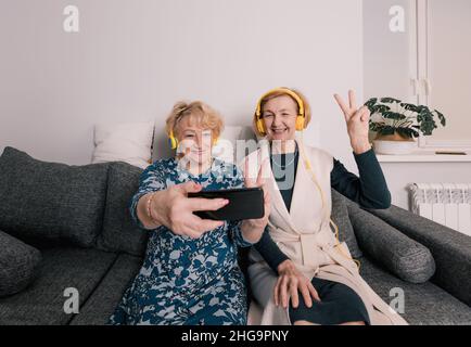 Zwei fröhliche, reife Großmütter in gelben Kopfhörern kommunizieren mit Blick auf den Smartphone-Bildschirm. Stockfoto