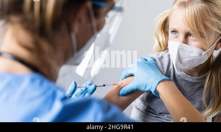 Blondes Mädchen, das den Arzt mit einer Spritze in der Hand ansieht, während es während einer Covid-19-Pandemie geimpft wurde oder eine Spritze erhielt Stockfoto