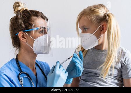 Blonde Mädchen in Gesichtsmaske im Impfzentrum werden geimpft. Spritze mit Impfstoff gegen Covid-19 Coronavirus, Grippe, gefährliche Infektionskrankheiten. Inje Stockfoto
