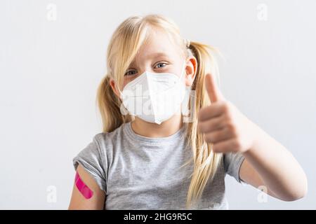 Blonde kleine Mädchen mit Gesichtsmaske geimpft gestikulierte Thumbs nach oben Armbinden zeigen, während sie geimpft wurden oder eine Spritze während covid-19 pand Stockfoto