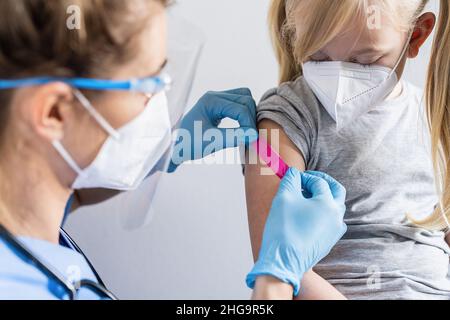 Arzt mit Schutzhandschuhen, der von einem blonden Mädchen eine Haftbandage auf einen Arm legt, nachdem er eine Impfung gegen das Covid-19-Coronavirus, Grippe, d erhalten hat Stockfoto