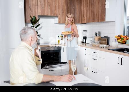 Lächelnde Frau hält Kuchen fast verschwommen Ehemann mit Champagner zu Hause Stockfoto