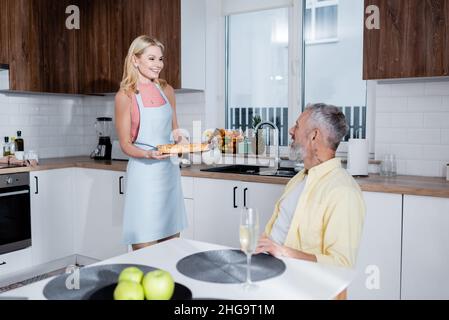 Positive Frau hält hausgemachten Kuchen in der Nähe des Mannes und Champagner auf dem Tisch in der Küche Stockfoto