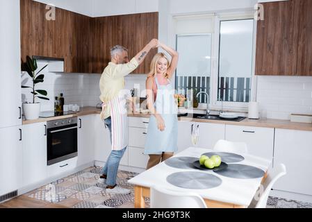Lächelnde Frau auf der Schürze, die mit ihrem Mann in der Nähe von Champagner in der Küche tanzt Stockfoto