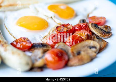 Frühstück mit Spiegeleiern, gegrillten Würstchen, gebratenen Pilzen und Kirschtomaten, Makroaufnahme auf einem weißen Teller. Stockfoto