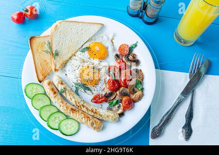 Frühstück mit Spiegeleiern, gegrillten Würstchen, gebratenen Pilzen und Kirschtomaten, frischen Gurkenscheiben und knusprigem Toast auf einem weißen Teller auf einem hölzernen BL Stockfoto