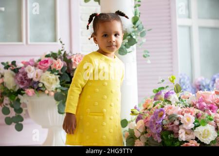 Kleine afroamerikanische Mädchen in einem gelben Kleid mit lockigen Zöpfen vor dem Hintergrund von schönen Blumensträußen. Stockfoto