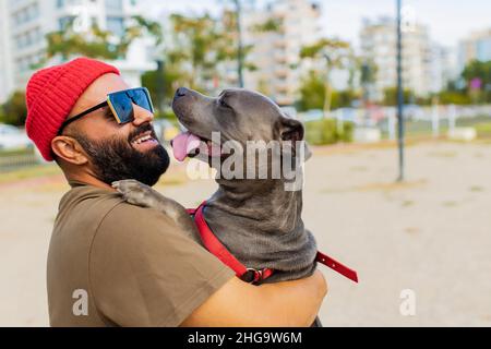 Porträt eines glücklichen Mannes in rotem Hut und Sonnenbrille mit amerikanischem Terrier in Hunde Walking Area Park in sity Stockfoto