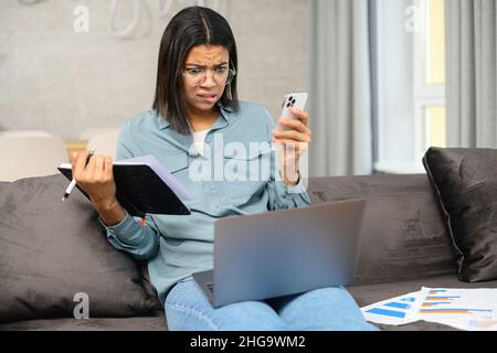 Konzentrierte, intelligente junge afroamerikanische Studentin in einer Brille, die auf der Couch mit einem Laptop auf dem Schoß sitzt und von zu Hause aus mit aufmerksamem Gesicht studiert Stockfoto