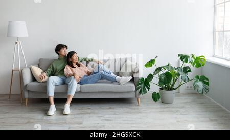 Positives junges asiatisches Paar, das sich auf dem Sofa entspannt, kuschelt und aus dem Fenster im Wohnzimmer blickt, freier Platz Stockfoto