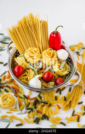 Gruppe ungekochter Pasta mit Gemüse in einem Edelstahlsieb auf weißer Oberfläche, vertikales Bild Stockfoto