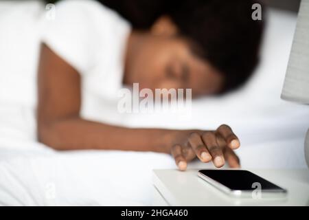 Schwarze Frau, die im Bett lag und den Wecker auf dem Handy abstellte Stockfoto