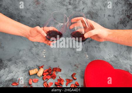 Zum Valentinstag zu Hause klirrende Rotweingläser anstoßen. Prost-Konzept mit Rotweingläsern und trockenen Rosenblättern. Spaß haben, gute Zeit zusammen. Stockfoto