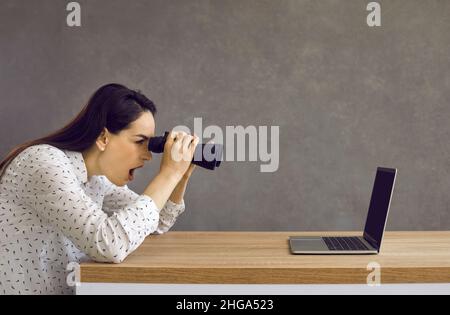 Lustig schockierte junge Frau, die Ferngläser hält und auf den Bildschirm eines Laptops schaut Stockfoto