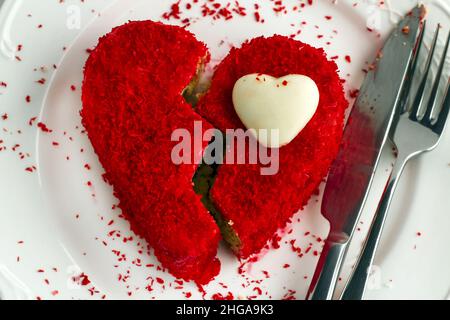 Süßer, roter, nasser Kuchen, halbiert wie gebrochenes Herz auf dem weißen Teller mit Messer und Gabel. Valentinstag-Konzept. Stockfoto