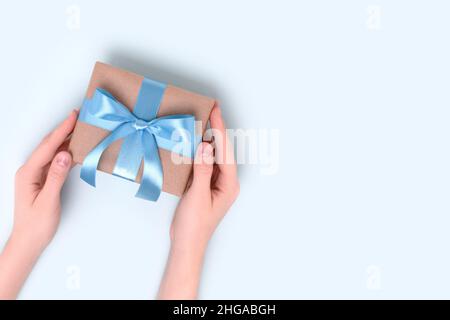 Weibliche Hände halten verpackte Geschenkbox auf einem blauen Hintergrund. Geschenk gebunden mit einem Band. Festliche Komposition mit Copyspace. Stockfoto