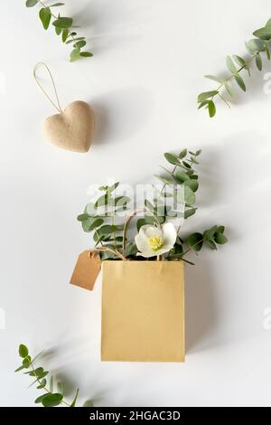 Basteltasche mit Eukalyptus im Winter und weißer Helleborus-Blume. Brauner Papieranhänger und Herz aus Pappe. Winter flach lag mit immergrünen Zweigen. Stockfoto