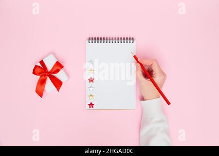 Weihnachten gemütlich Hintergrund. Weibliche Handschrift in leerem Notizbuch mit weißer Geschenkbox mit roter Schleife in pastellrosa. Planung des neuen Jahres, Ziele, To-Do-Liste oder Wunschliste-Konzept. Flach liegend, Kopierplatz Stockfoto