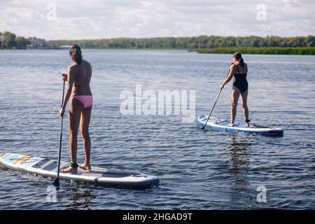 Zwei Frauen mittleren Alters pfützen beim Boarding auf dem wunderschönen blauen See mit Rudern in den Händen mit Bäumen und Schilf im Hintergrund, beide in Badeanzügen. Aktiv Stockfoto
