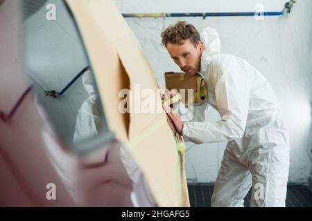 Auto Painter maskiert Karosserie mit Klebeband und Papier vor dem Lackieren. Mechaniker in einem weißen Schutzanzug arbeitet in einer Garage Stockfoto